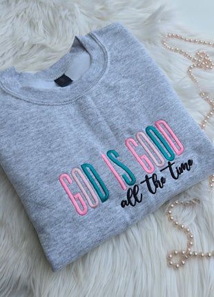 A folded heather gray sweatshirt with colorful embroidered text reading 'GOD IS GOOD all the time' in pink, teal, and black, styled on a white faux-fur surface with a pearl necklace nearby.