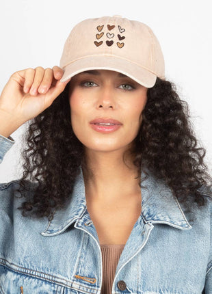 A woman wearing a light beige baseball cap with embroidered hearts, paired with a light blue denim jacket, against a plain white background.