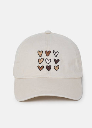 A light beige baseball cap featuring nine embroidered hearts in various shades of brown, beige, and white on the front panel, set against a plain light gray background.