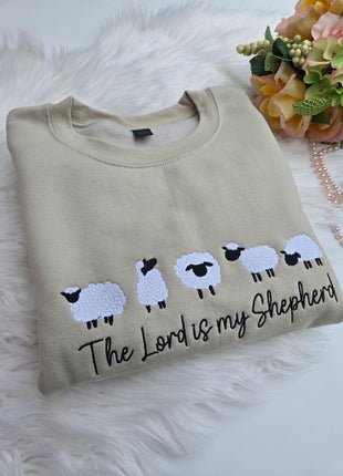 A folded beige sweatshirt with a graphic of five white sheep and the text 'The Lord is my Shepherd' in black, displayed on a white faux fur surface with decorative flowers and a pearl necklace.