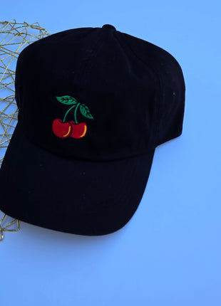 A black baseball cap featuring an embroidered cherry motif on the front, displayed on a solid blue background with a gold wire mesh prop on the left.