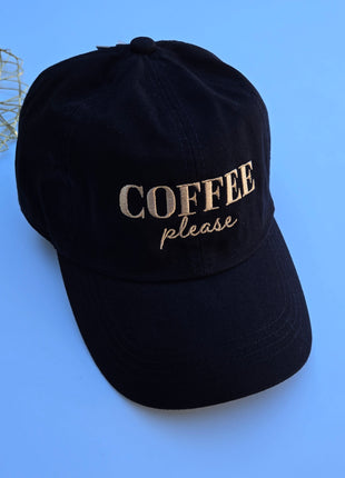 A black cotton baseball cap with 'COFFEE please' embroidered on the front in beige, set against a solid blue background with a decorative gold wire mesh in the top-left corner.