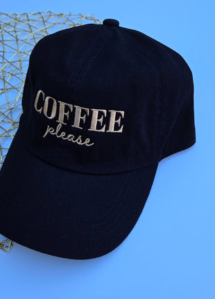 A black baseball cap with 'COFFEE please' embroidered on the front, styled on a bright blue background next to a gold wire decorative object.