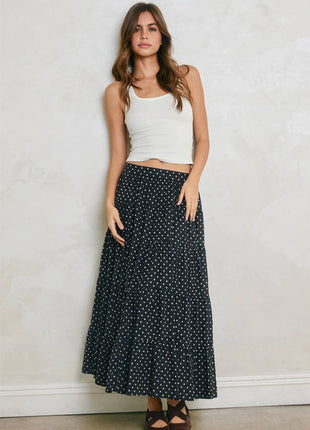 A woman modeling a long black maxi skirt with a white polka dot pattern, paired with a white ribbed tank top and brown sandals, standing against a textured off-white wall.