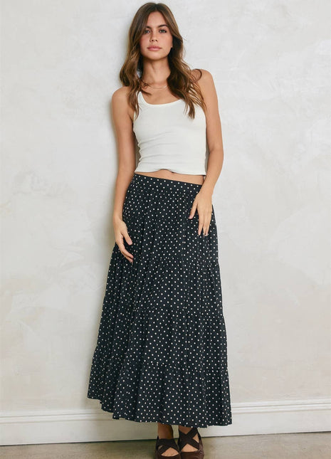 A woman modeling a long black maxi skirt with a white polka dot pattern, paired with a white ribbed tank top and brown sandals, standing against a textured off-white wall.