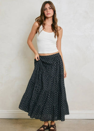 A woman modeling a long black skirt with a white polka dot pattern, paired with a white ribbed tank top and brown sandals, standing against a textured light gray wall.