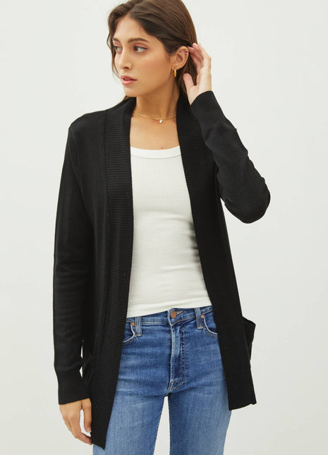 A woman wearing a black open-front knit cardigan over a white tank top and blue jeans, photographed against a plain light gray studio background.