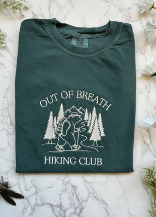 Folded green t-shirt with 'Out of Breath Hiking Club' design on a marble surface with flowers.
