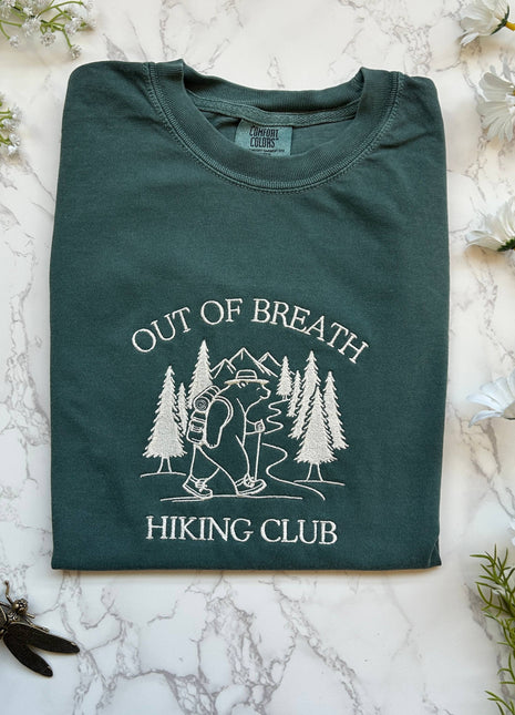 Folded green t-shirt with 'Out of Breath Hiking Club' design on a marble surface with flowers.
