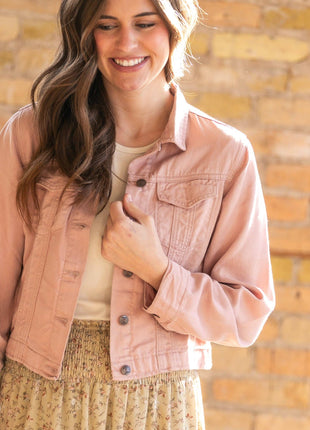 Woman wearing a pink jacket and floral skirt against a brick wall.