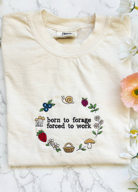 Folded cream-colored sweatshirt with embroidered design and text on a marble surface with flowers.