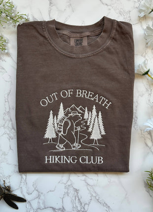 Folded brown t-shirt with 'Out of Breath Hiking Club' design on a marble surface with flowers.