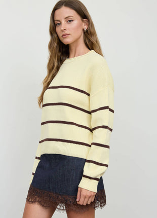 A woman modeling a pale yellow knit sweater with dark brown horizontal stripes, paired with a dark denim skirt featuring a brown lace hem, against a light gray studio background.