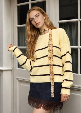 A woman models a cream and black striped knit sweater with a floral-patterned tie-neck detail, paired with a dark denim mini skirt with a lace hem, standing in front of a white paneled door.