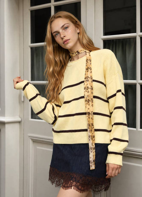 A woman models a cream and black striped knit sweater with a floral-patterned tie-neck detail, paired with a dark denim mini skirt with a lace hem, standing in front of a white paneled door.