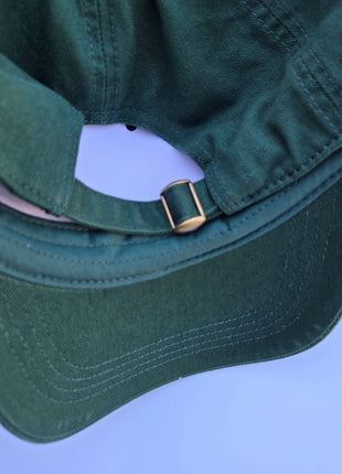 Close-up of the back of a dark green baseball cap with a curved brim and a small gold metal buckle on the strap, set against a light blue background.