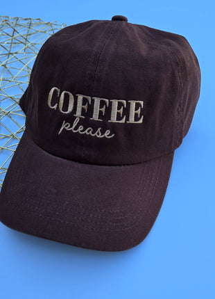 A dark brown cotton baseball cap with 'COFFEE please' embroidered on the front, set against a bright blue background with a gold wire mesh prop on the left.