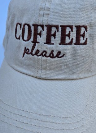 A close-up of a light beige cotton baseball cap with 'COFFEE please' embroidered on the front, set against a solid light blue background.
