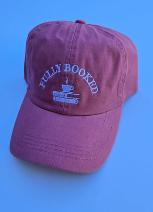 A dusty mauve-purple baseball cap with light blue embroidered text reading 'FULLY BOOKED' and a small book-and-tapestry graphic on the front, set against a solid blue background.