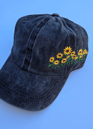 A dark washed denim baseball cap featuring embroidered yellow sunflowers with green stems on the side, set against a solid blue background.