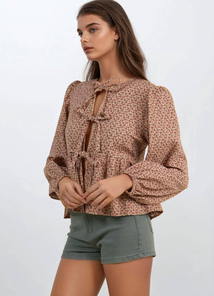 A woman modeling a long-sleeve blouse with a small-scale floral pattern in brown and beige, featuring a keyhole front with tie details, paired with olive green shorts against a plain light gray background.