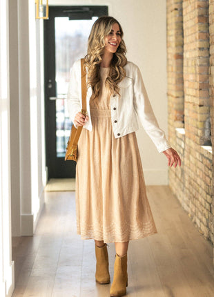 A woman stands in a hallway wearing a beige midi dress with a lace hem, a white button-up jacket, and tan ankle boots, while carrying a tan shoulder bag.