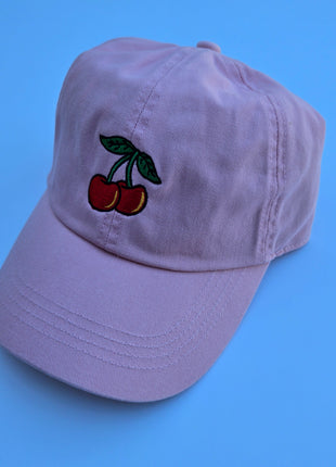 A light purple baseball cap featuring an embroidered design of two red cherries with green leaves on the front, set against a solid blue background.