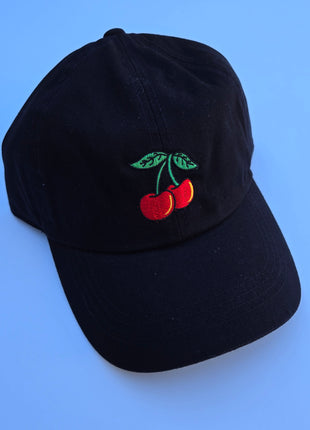 A black baseball cap featuring an embroidered design of two red cherries with green leaves on the front, set against a solid blue background.