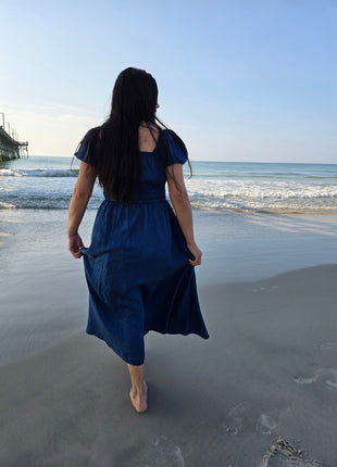A woman with long dark hair walks barefoot on a sandy beach, viewed from behind, wearing a dark blue midi dress with puff sleeves.
