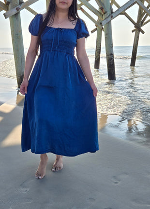 A woman wearing a royal blue midi dress with puff sleeves and a smocked bodice, standing on a sandy beach under a wooden pier structure with the ocean in the background.