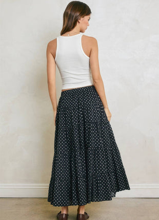 A woman shown from the back wearing a white ribbed tank top and a black midi skirt with a small white polka dot pattern, standing against a textured off-white wall.