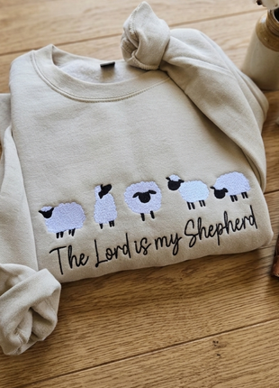 A folded cream-colored sweatshirt on a wooden tabletop, featuring embroidered sheep and the text 'The Lord is my Shepherd'