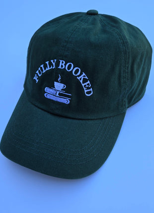 A dark forest green baseball cap with light blue embroidered text reading 'FULLY BOOKED' above an illustration of a coffee cup on stacked books, shown on a solid blue background.