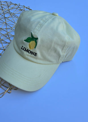 A pale yellow-green cotton baseball cap with an embroidered lemon and the word 'LIMONE' on the front, styled on a blue background next to a gold wire mesh tray.