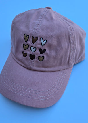 A dusty purple cotton baseball cap featuring embroidered hearts in various colors on the front, set against a solid blue background.