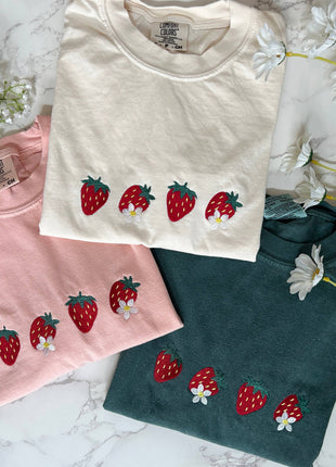 Three t-shirts in pink, white, and green with strawberry designs on a marble surface.