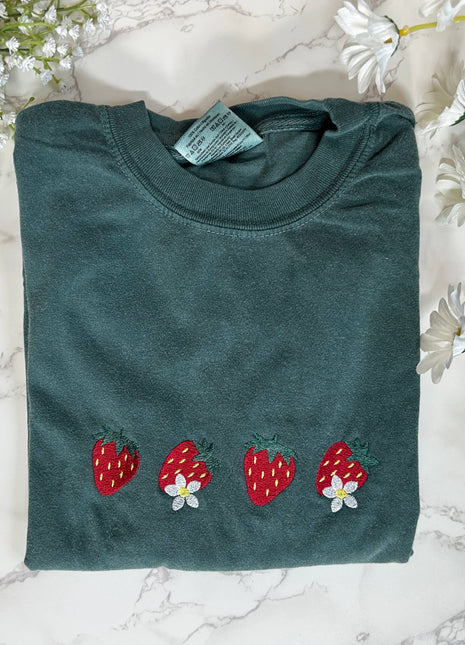 Folded teal shirt with strawberry designs on a marble surface with flowers.