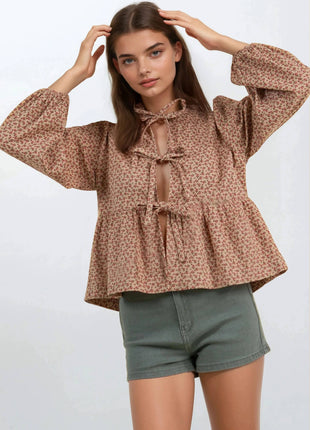 A woman modeling a brown and beige floral print long-sleeve blouse with a tie-neck detail, paired with olive green shorts, against a light gray studio background.