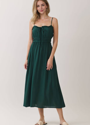 A woman modeling a dark green midi dress with thin spaghetti straps, a smocked bodice, and a gathered skirt, paired with beige slide sandals against a light beige studio background.