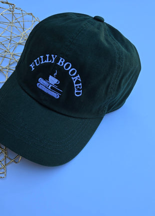 A dark forest green baseball cap with 'FULLY BOOKED' and a coffee cup graphic embroidered in light blue on the front, styled on a blue background next to a gold wire mesh sphere.