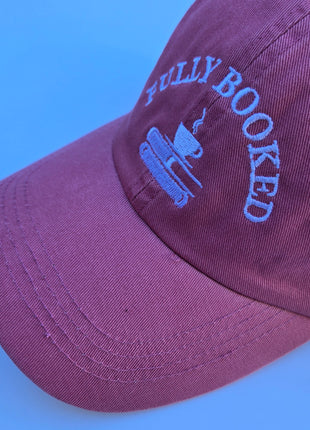A close-up of a dusty burgundy baseball cap with light blue embroidered text reading 'FULLY BOOKED' and a coffee cup graphic, set against a solid blue background.