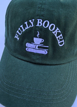 A dark green baseball cap with white embroidered text reading 'FULLY BOOKED' above a small coffee cup and books graphic, shown against a light blue background.