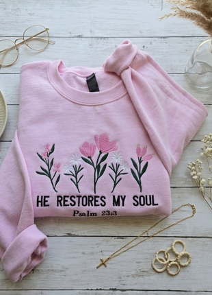 A light pink crewneck sweatshirt with a graphic of pink and white flowers and the text 'HE RESTORES MY SOUL Psalm 23:3' laid flat on a white wooden surface with decorative props including glasses, plates, a vase, and jewelry.