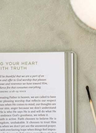 Open book with text on a surface next to a candle and small plant