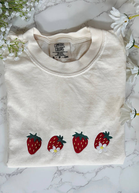 Folded white sweatshirt with strawberry designs on a marble surface with flowers.