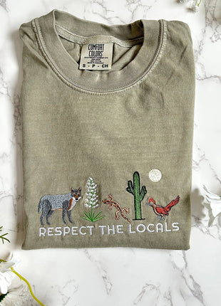 Folded green t-shirt with desert-themed graphics and 'Respect the Locals' text on a marble surface.