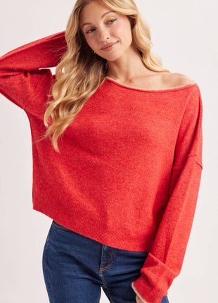 A woman wearing a bright red off-the-shoulder knit sweater and blue jeans, photographed against a clean light gray studio background.