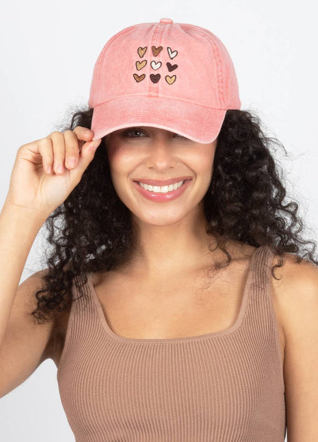 A smiling woman with curly dark hair wears a pink baseball cap with small embroidered hearts on the front, holding the brim with one hand against a light gray background.