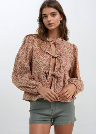 A woman modeling a long-sleeve blouse with a small-scale floral pattern in dusty pink and beige, featuring a keyhole tie neckline and ruffled front hem, paired with olive green denim shorts against a light gray studio background.