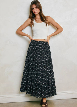 A woman modeling a long black maxi skirt with a white polka dot pattern, paired with a white ribbed tank top and brown sandals, standing against a textured off-white wall.